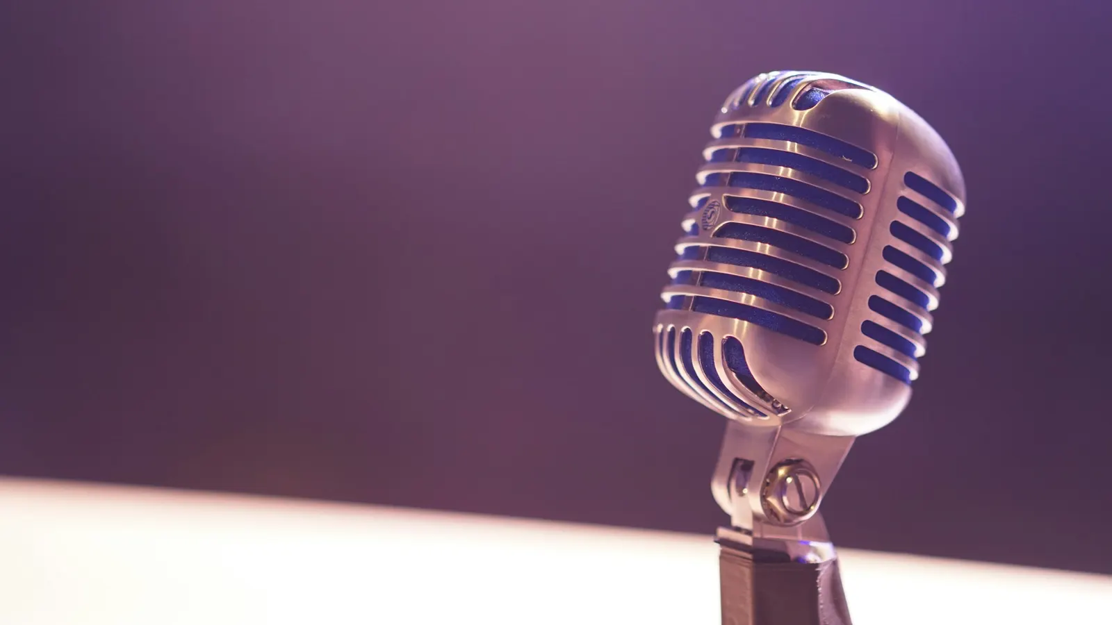 A broadcast microphone on a desk arm in a home office with a blurred monitor in the background
